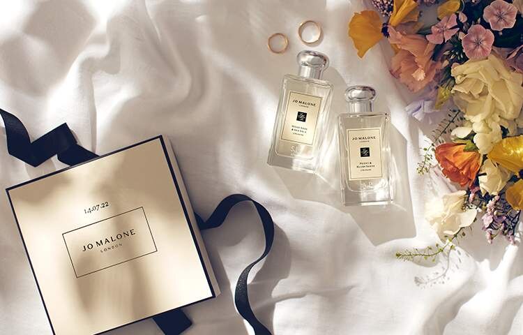 Image of two engraved jo malone colognes next to gold wedding rings & colourful flowers