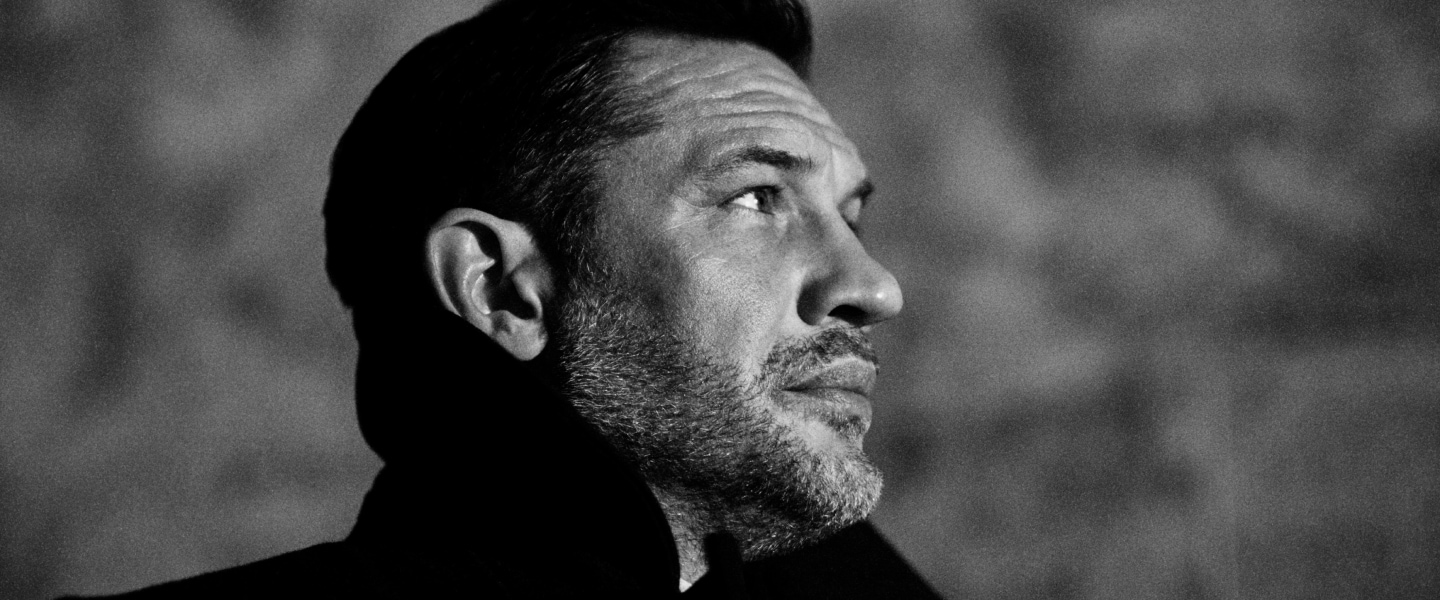 A side portrait of Tom Hardy in black and white, gazing into the distance. He wears a formal coat, with an upturned collar.