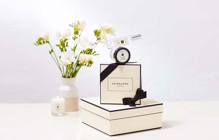 image of jo malone cream and black boxes stacked with a body creme balanced on top and flowers in a vase behind